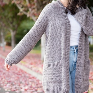 May include: A gray crocheted cardigan with pockets and a long, open front. The cardigan is worn over a white t-shirt and blue jeans.
