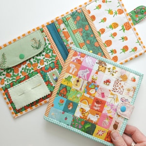 以下が含まれることがあります： パッチワークのデザインが施された布製の針刺しケースが2つ。1つのケースは緑とオレンジの花柄で、小さなポケットが付いています。もう1つのケースは、鳥、猫、ギターなど、さまざまな小さな画像が描かれたカラフルなパッチワークのデザインです。パッチワークケースには、「My friends are my happy place」というテキストが表示されています。