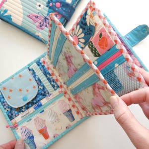 K&ouml;nnte beinhalten: Gesteppte Stoffbrieftasche mit blauem und wei&szlig;em Schmetterlingsmuster, ge&ouml;ffnet, um mehrere F&auml;cher freizugeben. Mit Rei&szlig;verschlusstasche mit Eiswaffel-Grafiken und einem kleinen M&uuml;nzfach mit Sternakzenten. Der Geldbeutel hat einen Druckknopfverschluss.