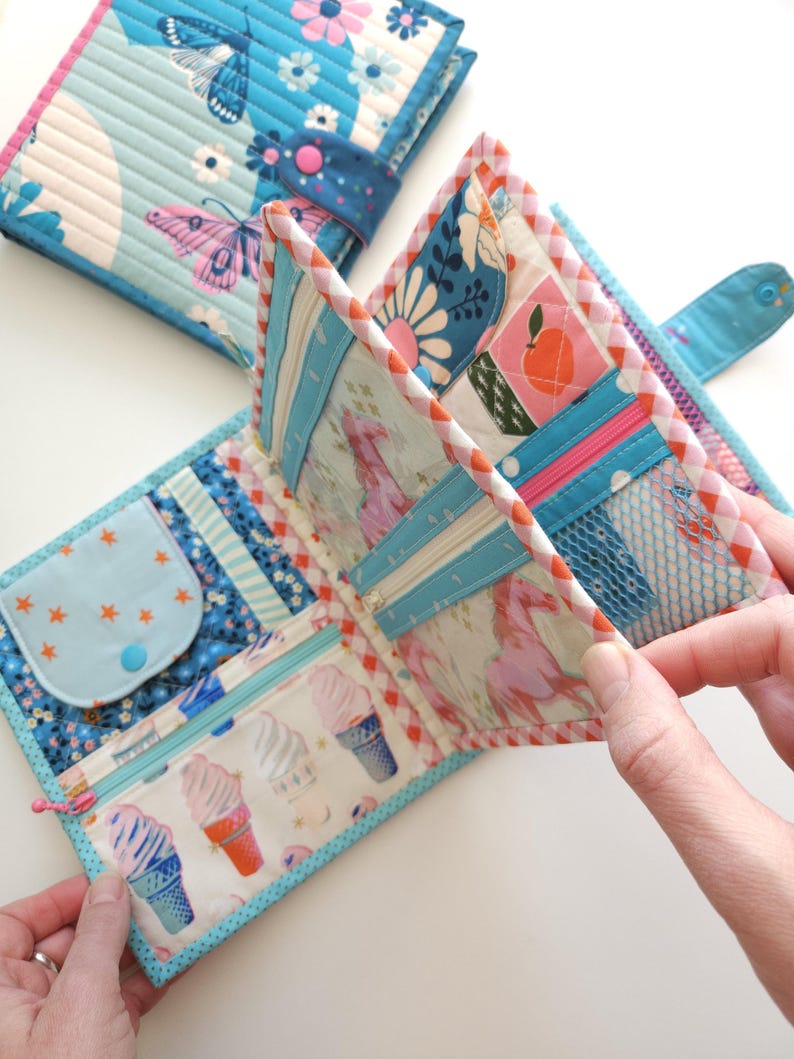 K&ouml;nnte beinhalten: Gesteppte Stoffbrieftasche mit blauem und wei&szlig;em Schmetterlingsmuster, ge&ouml;ffnet, um mehrere F&auml;cher freizugeben. Mit Rei&szlig;verschlusstasche mit Eiswaffel-Grafiken und einem kleinen M&uuml;nzfach mit Sternakzenten. Der Geldbeutel hat einen Druckknopfverschluss.