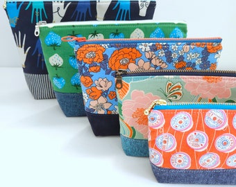 May include: Five colourful zippered pouches with different patterns. The pouches are made of fabric and have a denim trim. The patterns include floral, geometric, and abstract designs.
