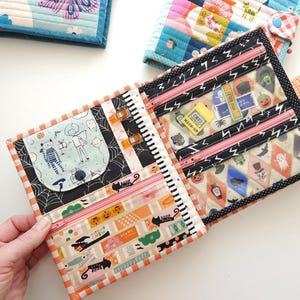 K&ouml;nnte beinhalten: Quilt-Geldb&ouml;rsen mit verschiedenen Mustern. Eine Geldb&ouml;rse ist ge&ouml;ffnet und zeigt mehrere Rei&szlig;verschlussf&auml;cher und einen Druckknopfverschluss. Die offene Geldb&ouml;rse hat ein Halloween-Design mit orangefarbenen, schwarzen und wei&szlig;en Akzenten.