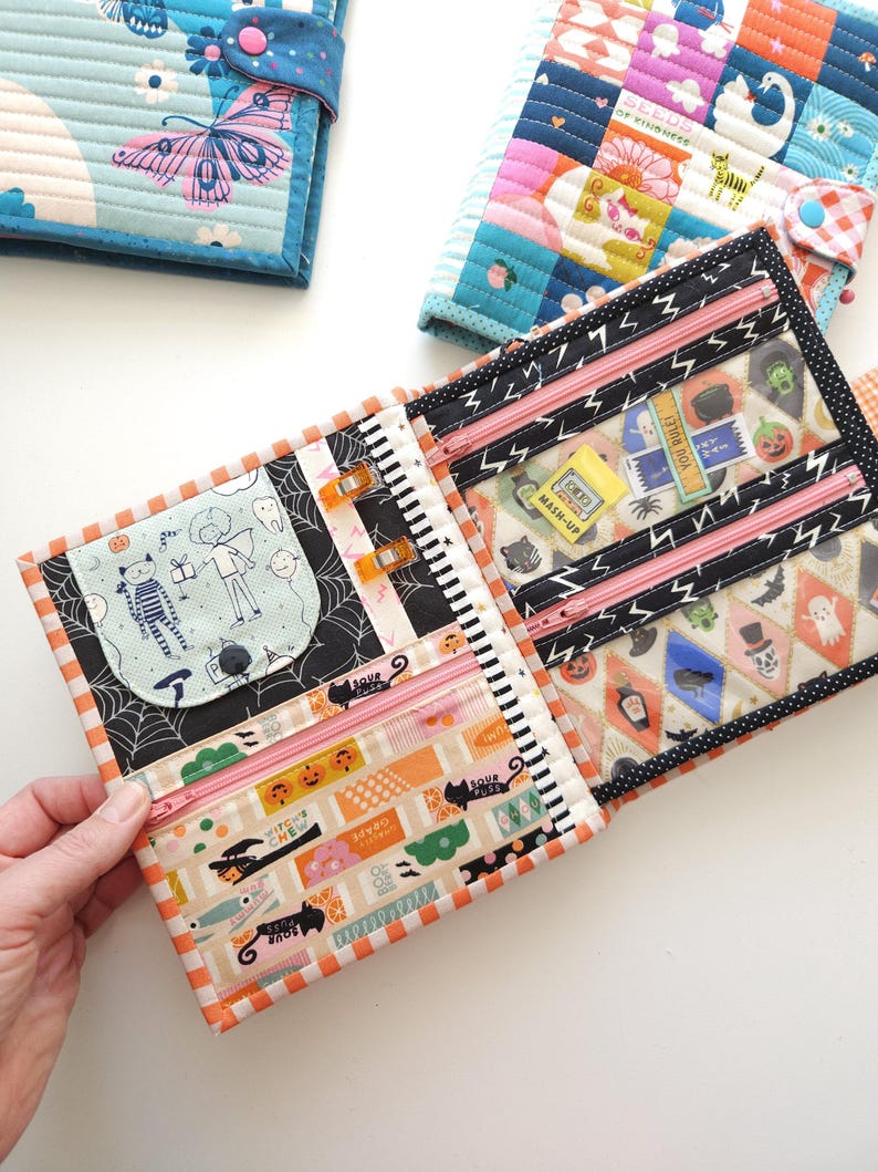 K&ouml;nnte beinhalten: Quilt-Geldb&ouml;rsen mit verschiedenen Mustern. Eine Geldb&ouml;rse ist ge&ouml;ffnet und zeigt mehrere Rei&szlig;verschlussf&auml;cher und einen Druckknopfverschluss. Die offene Geldb&ouml;rse hat ein Halloween-Design mit orangefarbenen, schwarzen und wei&szlig;en Akzenten.