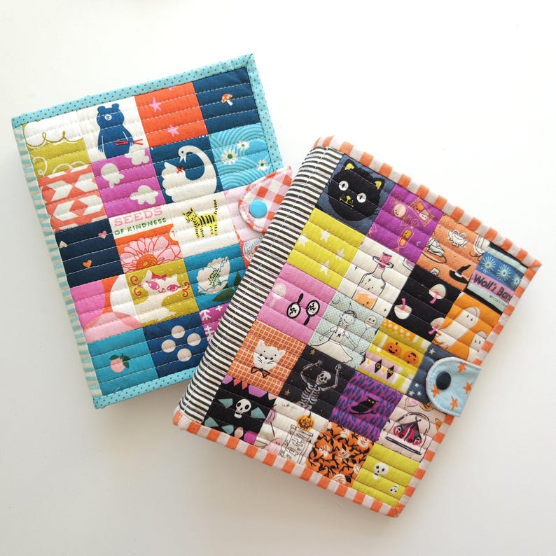 K&ouml;nnte beinhalten: Zwei bunte, gesteppte Stoffbuchh&uuml;llen. Eine zeigt einen blauen B&auml;ren, einen Schwan und den Text "SEEDS OF KINDNESS". Die andere hat eine schwarze Katze, ein Skelett und Halloween-Motive. Beide haben Knopfverschl&uuml;sse.