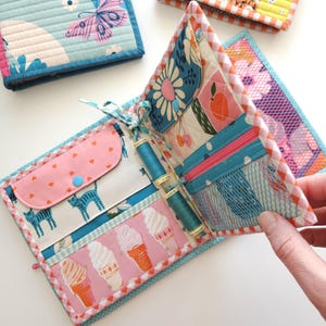 May include: Quilted fabric wallet with a variety of colorful patterns, including butterflies, flowers, and ice cream cones. The wallet is open, revealing pockets and spools of thread. The design features pink, blue, and white accents.