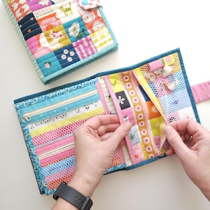 K&ouml;nnte beinhalten: Eine farbenfrohe, gesteppte Geldb&ouml;rse mit mehreren F&auml;chern, darunter Rei&szlig;verschlusstaschen und Netzteile. Die Geldb&ouml;rse hat ein Patchwork-Design mit verschiedenen Stoffmustern und -farben, darunter rosa, blau, gelb und Blumendrucke. Der Text "SEEDS OF KINDNESS" ist sichtbar.