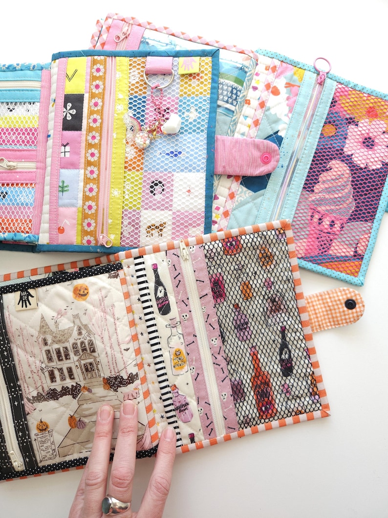 K&ouml;nnte beinhalten: Eine Sammlung farbenfroher, gesteppter Stoffbeutel mit Rei&szlig;verschl&uuml;ssen und Netztaschen. Zu den Designs geh&ouml;ren Blumenmuster, Halloween-Motive mit einem Spukhaus und dekorative Streifen. Einige Beutel haben Druckkn&ouml;pfe und Anh&auml;nger.