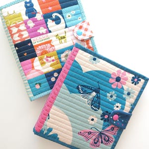 K&ouml;nnte beinhalten: Zwei farbenfrohe, gesteppte Stoffbuchh&uuml;llen. Eine zeigt ein Patchwork-Design mit einem B&auml;ren, einem Schwan und dem Text "SEEDS OF KINDNESS". Die andere hat Schmetterlinge und Blumen. Beide haben Knopfverschl&uuml;sse und sind mit passenden Stoffen eingefasst.