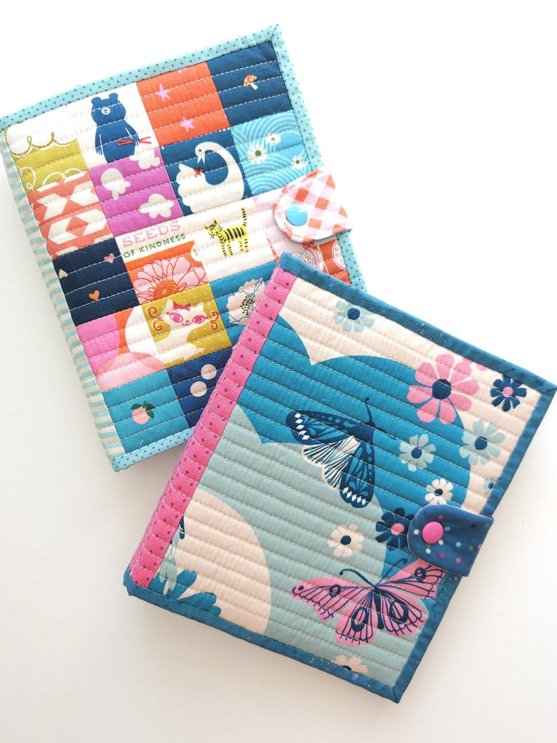 K&ouml;nnte beinhalten: Zwei farbenfrohe, gesteppte Stoffbuchh&uuml;llen. Eine zeigt ein Patchwork-Design mit einem B&auml;ren, einem Schwan und dem Text "SEEDS OF KINDNESS". Die andere hat Schmetterlinge und Blumen. Beide haben Knopfverschl&uuml;sse und sind mit passenden Stoffen eingefasst.