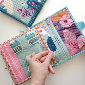 K&ouml;nnte beinhalten: Quilt-Organizer aus Stoff mit einem Schmetterlings- und Blumenmuster. Der Organizer hat mehrere F&auml;cher, darunter Rei&szlig;verschlusstaschen. Das Farbschema umfasst Blau-, Rosa- und Wei&szlig;t&ouml;ne. Der Organizer ist f&uuml;r die Aufbewahrung kleiner Gegenst&auml;nde konzipiert.