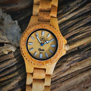 May include: A wooden wristwatch with a light brown bamboo band and a round face. The face has white markings and a blue second hand. The watch face says "Nalu Quartz".