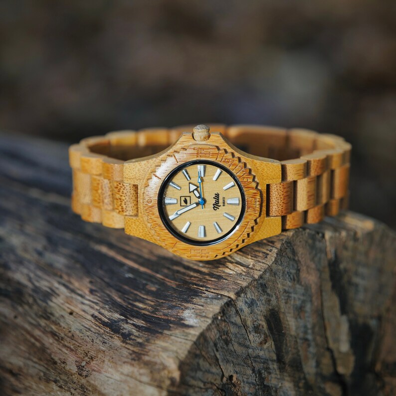 May include: A wooden wristwatch with a light brown bamboo band and a cream-colored face. The watch face has black markings and a blue second hand. The watch face also has the word "Nalu" printed on it.