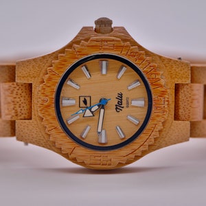 May include: A wooden wristwatch with a light brown bamboo band and a cream-colored face. The watch face has black markings and a blue second hand. The watch face also has the word "Malu" printed on it.