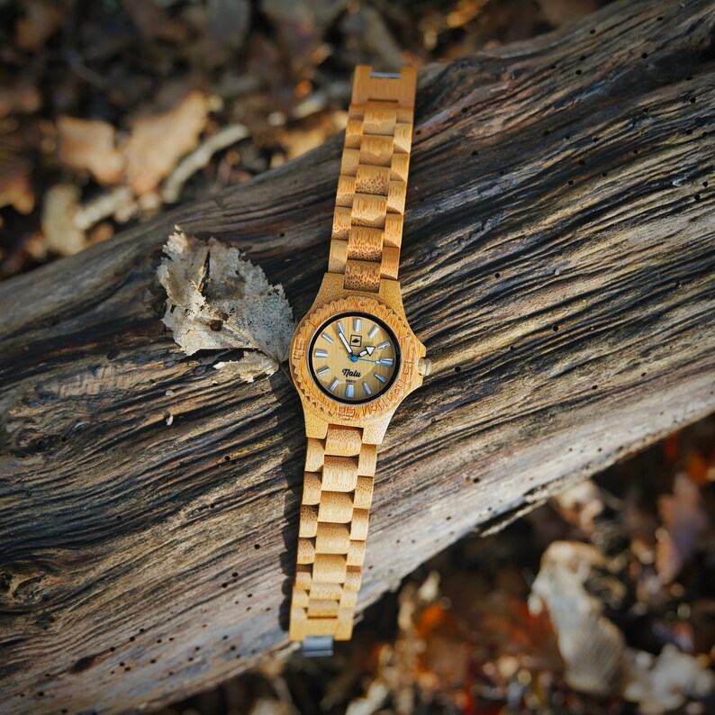 May include: A wooden wristwatch with a light brown band and a round, white face with black markings. The watch is on a brown wooden surface.