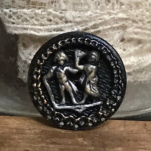 May include: A black button with a silver design of two figures dancing. The button has a decorative border.