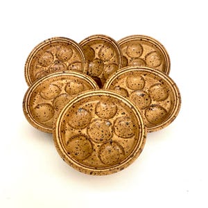 May include: A collection of seven speckled, tan-colored ceramic escargot dishes. Each dish has seven circular indentations for holding snails, with a raised rim. The dishes are arranged in a circular pattern against a white background.