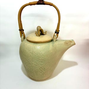 May include: A light green ceramic teapot with a textured surface and a leaf pattern. It features a bamboo handle and a lid with a decorative knob. The spout is angled, and the teapot is designed for pouring.