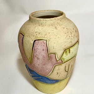 May include: A beige ceramic vase with a textured surface and a wide opening. The vase features a colorful geometric design with pink, blue, and yellow shapes. The vase is approximately 8 inches tall.