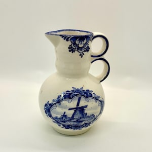 May include: A white ceramic pitcher featuring blue hand-painted floral patterns around the rim and a central oval medallion depicting a windmill landscape. The vessel has a rounded body, a narrow neck, and two stacked circular handles on the side.
