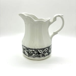 May include: White ceramic pitcher with a decorative black and white floral pattern around the base. The pitcher has a curved handle and a scalloped rim. A classic piece for serving beverages.