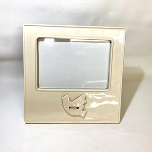 May include: Cream-colored ceramic picture frame with a rectangular opening. A raised dove design with a button detail is on the lower right corner. The frame is square and has a glossy finish.