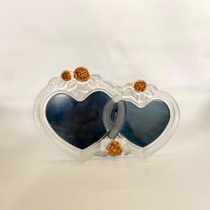 May include: A clear glass double heart-shaped picture frame. The frame has a frosted finish with amber-colored floral accents. The two hearts are interlocked, creating a romantic and decorative piece.