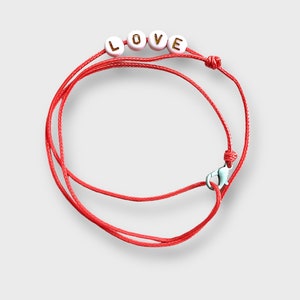 May include: Red string bracelet with white beads that spell out the word "LOVE" in gold lettering.