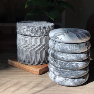 May include: Two decorative containers made of gray and white marbled resin. One container is a cylinder with a lid and the other is a stack of five round discs.