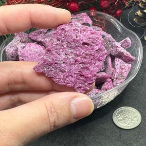 May include: A close-up of a small bowl filled with pink ruby rough crystals. The crystals are irregular in shape and have a rough, textured surface. A hand is holding a large piece of the ruby rough.