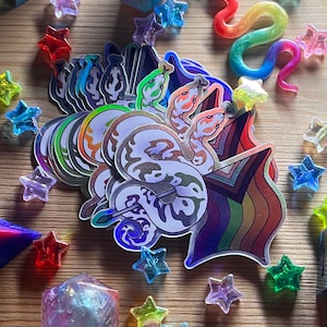 Könnte beinhalten: Eine Sammlung von Aufklebern, Würfeln und sternförmigen Anhängern. Die Aufkleber zeigen ein Schlangenmuster mit Regenbogen- und schillernden Akzenten. Die Würfel sind mehrseitig und durchscheinend. Die Sternanhänger sind in verschiedenen Farben.