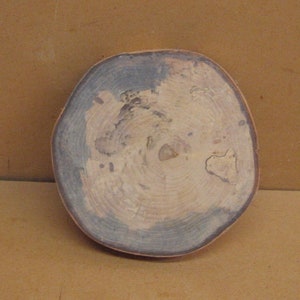 May include: A round, wooden coaster with a natural edge. The coaster features concentric rings in shades of beige, gray, and blue. The wood grain is visible, adding to the rustic aesthetic. The coaster is approximately 4 inches in diameter.