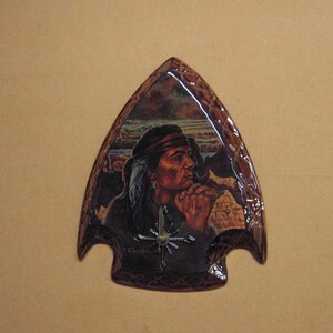 May include: A brown arrow-shaped plaque with a painted image of a Native American man in a feathered headdress, looking to the side. The plaque has a brown wooden frame.