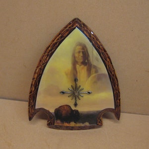 May include: A wooden arrowhead-shaped wall clock with a Native American image and a buffalo in the background. The clock face has a starburst design.