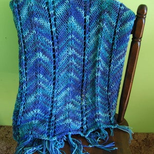 May include: A blue and teal knitted throw blanket with a chevron pattern and fringe. The blanket is draped over a wooden chair.