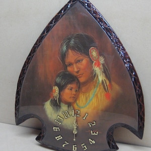May include: A wooden clock shaped like an arrowhead with a painting of a Native American mother and daughter. The clock face has Roman numerals and a gold-colored hand.