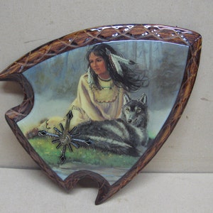 May include: A wooden plaque with a painted image of a Native American woman with a wolf. The woman is wearing a feathered headdress and a beaded necklace. The wolf is lying down next to her. The plaque is shaped like a teardrop and has a brown wooden frame.