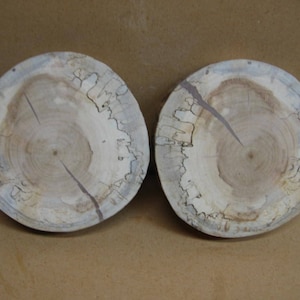 May include: Two round wooden plates with a natural wood grain pattern and a light brown color. The plates have a slightly raised edge and a smooth surface.