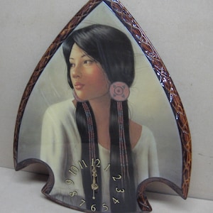 May include: A wooden arrowhead-shaped clock with a picture of a Native American woman with long black hair and a pink and blue beaded necklace. The clock face has Roman numerals and a gold-colored hand.