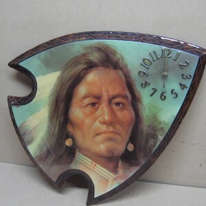 May include: A wooden clock shaped like an arrowhead with a picture of a Native American man on the face. The clock face has Roman numerals and a silver hand.