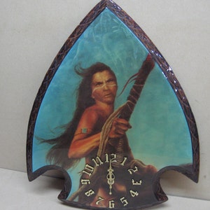 May include: A wooden clock shaped like an arrowhead with a Native American woman holding a spear in front of a blue and white background. The clock face has Roman numerals and a gold-colored hand.