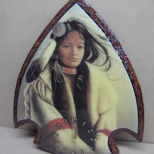 May include: A wooden clock shaped like an arrowhead with a picture of a Native American woman in a white and brown feathered headdress. The clock face has Roman numerals and a gold-colored trim.