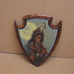 May include: Arrowhead-shaped wall art featuring a Native American man playing a flute, with a wolf howling in the background under a full moon. The artwork has a decorative, woven-style border in shades of brown and is set against a neutral background.