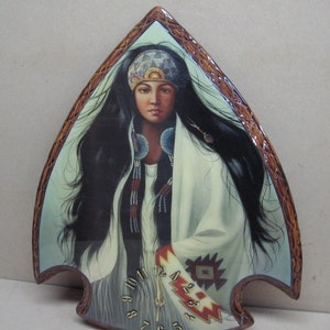 May include: A wooden clock shaped like a Native American arrowhead with a painted image of a woman with long black hair wearing a white shawl and a beaded necklace. The clock face is visible with Roman numerals and a gold hand.