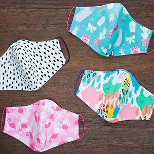 May include: Four different fabric face masks with various patterns. The masks are all folded in half and have elastic ear loops. The patterns include black and white polka dots, pink flamingos, turquoise with a variety of colorful illustrations, and a colorful abstract pattern.