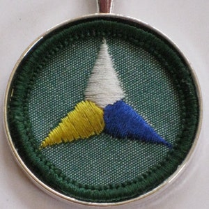 Puede incluir: Un collar con un colgante de plata y un c&iacute;rculo de tela verde con un tri&aacute;ngulo bordado en blanco, amarillo y azul.