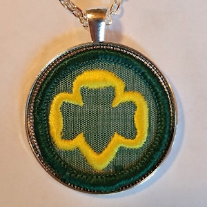 May include: A silver chain necklace with a round pendant featuring a green and yellow embroidered Girl Scout trefoil.