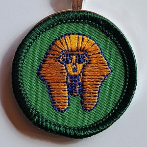 May include: A round pendant necklace featuring an embroidered design of an Egyptian pharaoh's head. The pendant has a green background with orange and blue embroidery, and a silver chain.
