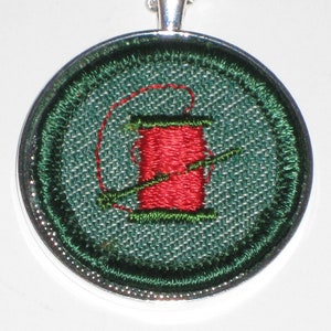 May include: A silver pendant necklace with a green and red embroidered spool of thread design.