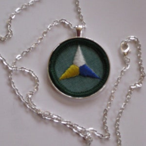 Puede incluir: Un collar de cadena de plata con un colgante redondo con fondo verde y un tri&aacute;ngulo blanco, amarillo y azul.