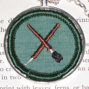 May include: A silver pendant necklace with a green fabric circle featuring two crossed paintbrushes. The paintbrushes are brown and black.